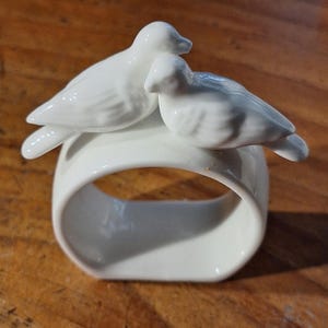 May include: A white ceramic napkin ring featuring two sculpted birds perched side by side on the top edge. The birds have smooth, glossy surfaces with subtle moulded wing details, and the ring itself has a rounded, rectangular opening.