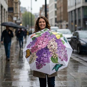 May include: White umbrella featuring a floral design with purple, pink, and white flowers, and butterflies. The umbrella is open, showcasing the colorful artwork. The background shows a rainy street scene with people and cars.