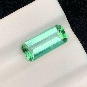 May include: A rectangular, emerald-cut gemstone with a vibrant green hue. The gem is displayed on a white surface against a dark background. The gemstone's facets reflect light, showcasing its clarity and brilliance. This item is suitable for jewelry making.