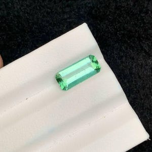 May include: A rectangular, emerald-cut, light green gemstone. The gem is displayed on a white surface against a black background. The gemstone's facets reflect light, showcasing its clarity and color. This item is suitable for jewelry making.