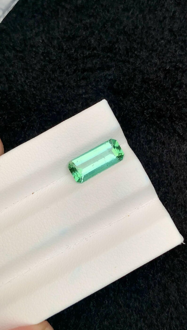 May include: A rectangular, emerald-cut, light green gemstone. The gem is displayed on a white surface against a black background. The gemstone's facets reflect light, showcasing its clarity and color. This item is suitable for jewelry making.