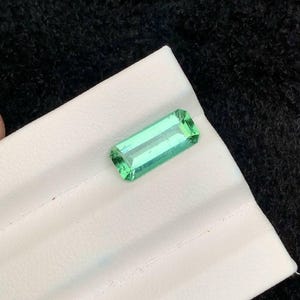 May include: A rectangular, faceted, light green gemstone. The gem is set on a white surface against a black background. The gemstone's cut allows light to pass through, highlighting its clarity and color.