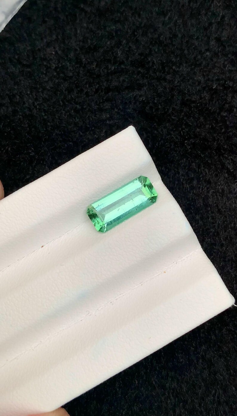 May include: A rectangular, faceted, light green gemstone. The gem is set on a white surface against a black background. The gemstone's cut allows light to pass through, highlighting its clarity and color.