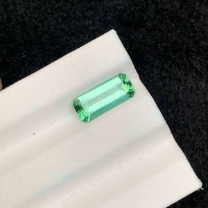 May include: A rectangular, light green gemstone with a polished surface. The gem is set on a white surface against a black background. The gemstone's shape is a long rectangle with cut corners.