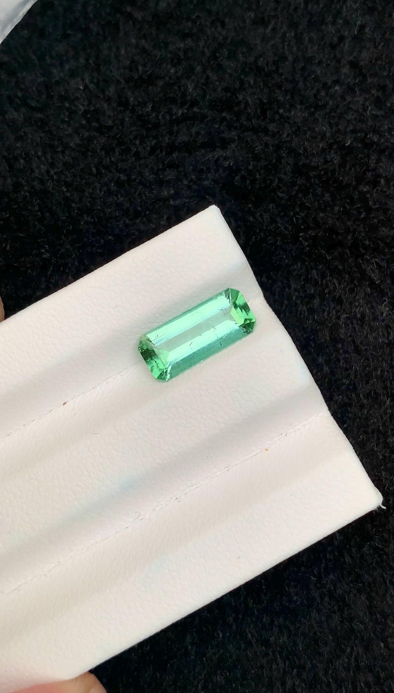 May include: A rectangular, light green gemstone with a polished surface. The gem is set on a white surface against a black background. The gemstone's shape is a long rectangle with cut corners.