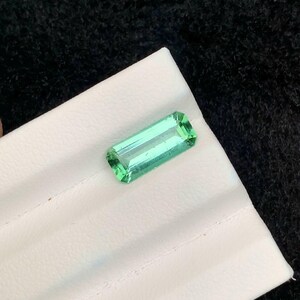 May include: A rectangular, light green gemstone with a faceted cut, resting on a white surface. The gem has a translucent quality, and the facets catch the light, creating a sparkling effect. The background is a dark, textured fabric.