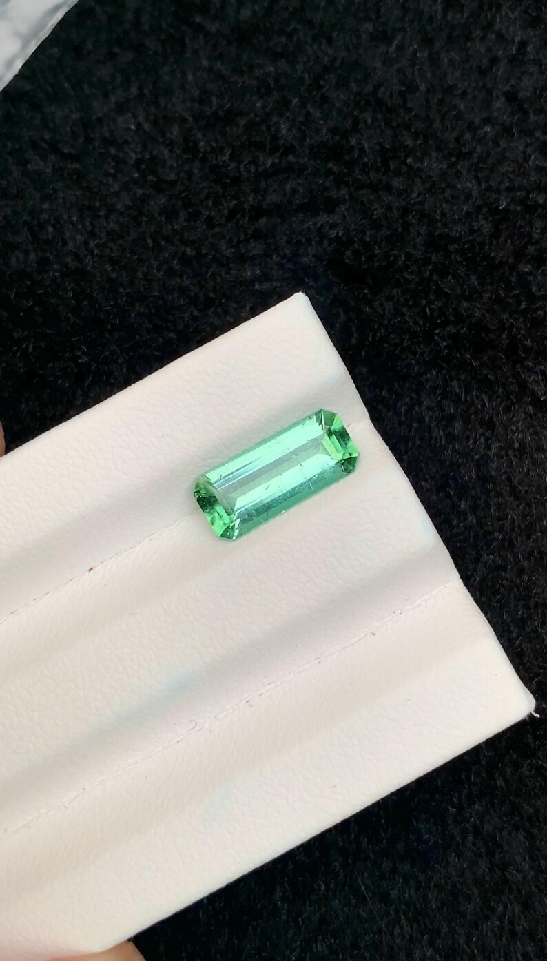 May include: A rectangular, light green gemstone with a faceted cut, resting on a white surface. The gem has a translucent quality, and the facets catch the light, creating a sparkling effect. The background is a dark, textured fabric.