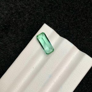 May include: A rectangular, emerald-green gemstone with a faceted cut. The gem is displayed on a white, textured surface against a dark background. The stone's clarity and color are prominent.
