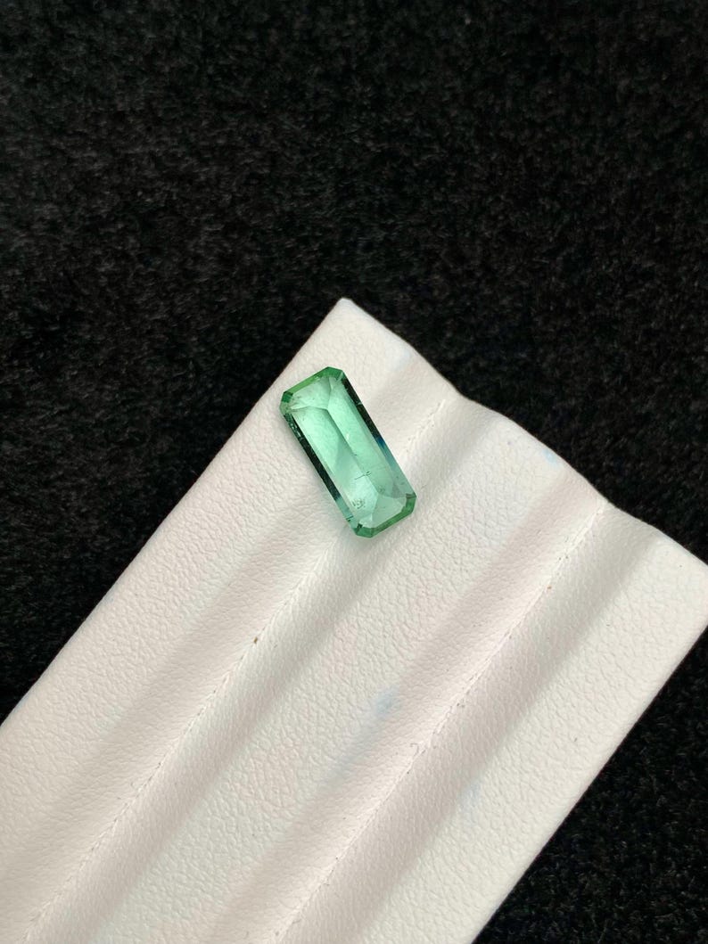 May include: A rectangular, emerald-green gemstone with a faceted cut. The gem is displayed on a white, textured surface against a dark background. The stone's clarity and color are prominent.
