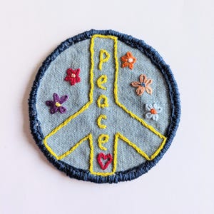 May include: Embroidered peace sign patch with the word "peace" in yellow thread. The patch is round with a blue denim background and a dark blue stitched border. Small embroidered flowers in various colours surround the peace symbol.