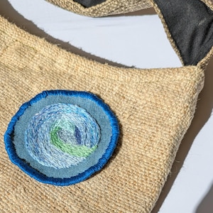 May include: A beige burlap shoulder bag with a dark grey strap. The bag has a circular embroidered patch with a blue, green, and white swirl design. The patch is outlined with a bright blue border.