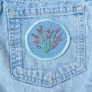 May include: A close-up of a light blue denim pocket with a circular embroidered patch. The patch has a light blue base and a floral design in pink, red, and green. It is edged with a light blue border.