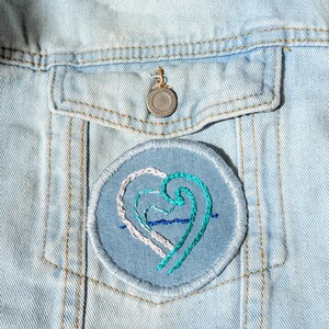 May include: A light blue denim jacket with a round patch featuring a heart design. The patch has a blue background with white and teal heart shapes and blue wave details. The jacket has a silver button.