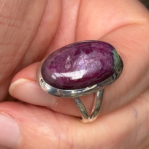 May include: A silver ring with a large, oval gemstone. The gemstone is a deep, purplish-red colour with a textured surface. The ring has a simple, split-shank design.