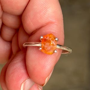 May include: A silver ring featuring a raw, orange-colored gemstone. The stone is irregularly shaped and held in place by four silver prongs. The ring band is thin and polished.