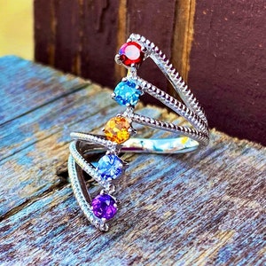 May include: A silver ring with a cascading design of six colorful gemstones. The stones are red, blue, orange, blue, purple, and red. The ring is set on a wooden surface.