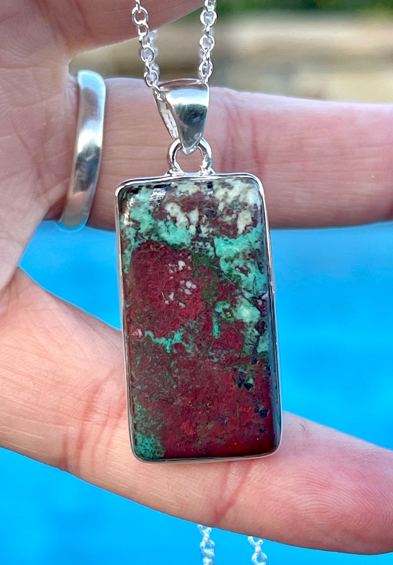 May include: A silver chain necklace with a rectangular pendant featuring a green and red gemstone. The pendant has a mottled pattern of green, red, and white.