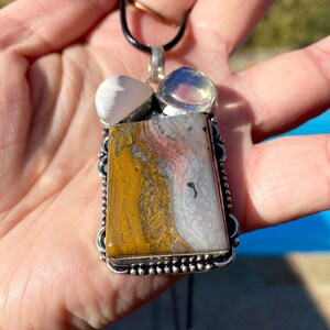 May include: A silver pendant necklace featuring a large rectangular yellow, white and pink agate stone. The pendant is surrounded by a silver border with small beads. The necklace is on a black cord.