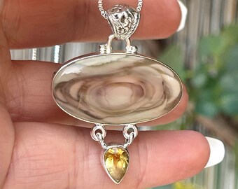 May include: A silver pendant necklace featuring an oval-shaped, cream-colored stone with brown swirls. Below the oval stone is a pear-shaped, yellow gemstone. The necklace is on a silver chain.