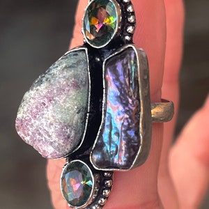 Könnte beinhalten: Ein Silberring mit einem großen, unregelmäßig geformten, hellvioletten Stein, einem rechteckigen, irisierenden Stein und zwei kleineren, runden, facettierten, irisierenden Steinen. Der Ring hat ein einzigartiges, künstlerisches Design.
