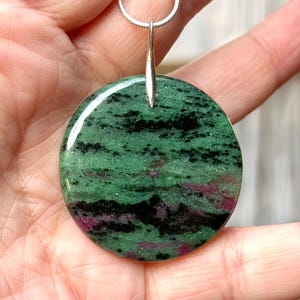 May include: A round pendant necklace featuring a polished green stone with black and red inclusions. The stone is suspended from a silver-colored chain. The pendant is a natural stone with a unique pattern.