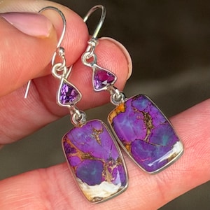 May include: Silver earrings with rectangular purple stones featuring gold and white veining. Each earring has a small, triangular purple stone above the larger stone. The earrings are held up by silver hooks.