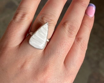 May include: A white, triangular ring with a textured surface. The ring is worn on a hand with pink and purple nail polish.