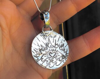 Op de afbeelding: Een zilveren ketting met een ronde hanger met een hart met vleugels. De hanger is getextureerd en heeft een gedetailleerd ontwerp.