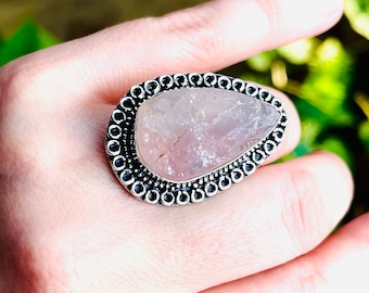 Pode incluir: Anel prateado com uma grande pedra de quartzo rosa em forma de gota. A pedra rosa claro está engastada numa moldura de metal prateado com uma borda decorativa de contas. O anel é mostrado numa mão, destacando o design e o tamanho.