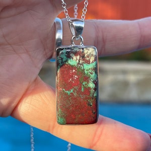 May include: A silver chain necklace with a rectangular pendant featuring a green and red stone. The stone has a mottled pattern.