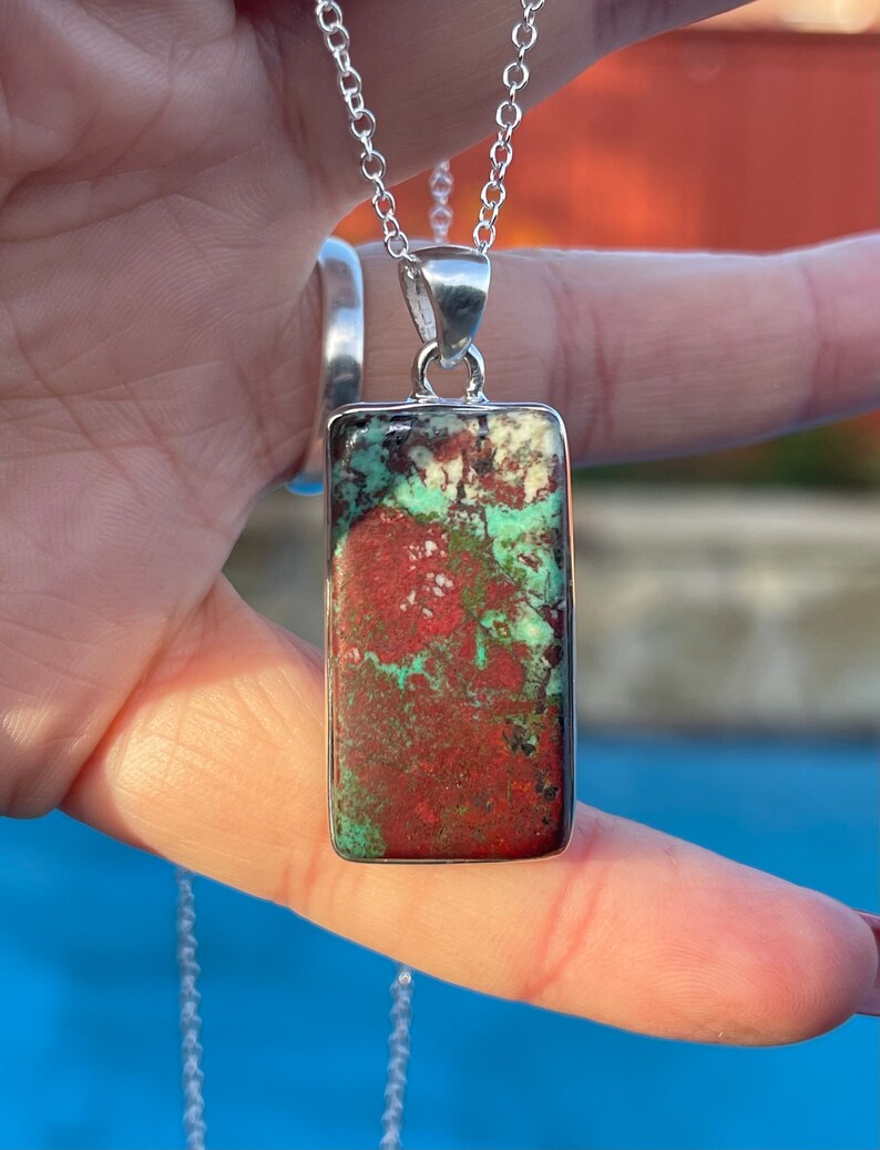 May include: A silver chain necklace with a rectangular pendant featuring a green and red stone. The stone has a mottled pattern.