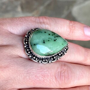 Op de afbeelding: Een zilveren ring met een grote, traanvormige groene edelsteen. De edelsteen heeft een gevlekt patroon en is gezet in een decoratieve zilveren band.