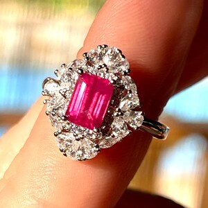 May include: A silver ring with a large, bright pink, emerald-cut gemstone surrounded by smaller, clear gemstones.