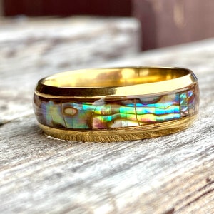 May include: A gold-toned ring with an iridescent abalone inlay. The ring features a band of colorful, shimmering abalone shell set within a gold frame. The ring is on a weathered wooden surface.