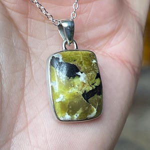 May include: A silver pendant necklace featuring a rectangular, olive-green stone with black and white inclusions. The stone is set in a silver bezel and hangs from a delicate silver chain. The pendant has a polished finish.