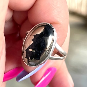 May include: A silver ring with an oval-shaped black and silver gemstone. The ring is set in a simple band.