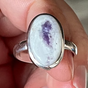 Può includere: Anello ovale con fascia argentata. L'anello presenta una pietra bianca con un disegno viola al centro. L'anello è tenuto dalle dita di una persona.