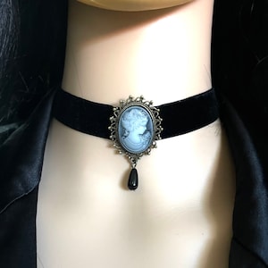 May include: A black velvet choker necklace with a silver cameo pendant featuring a woman's profile. The pendant has a black teardrop-shaped bead hanging from it.