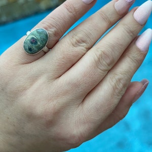 May include: A silver ring featuring a green and pink oval gemstone. The ring is worn on a finger with pink nail polish.