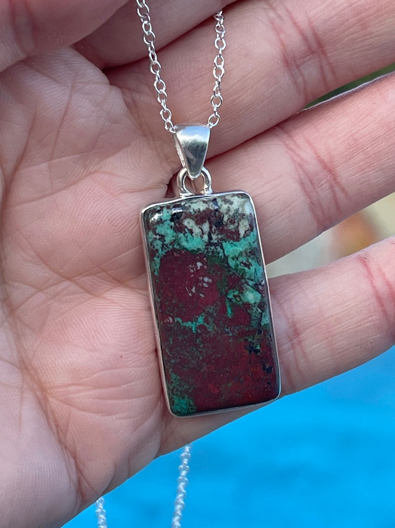 May include: A silver chain necklace with a rectangular pendant featuring a green, red, and white patterned stone.