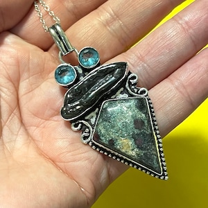 May include: A silver pendant necklace with a green and black stone, accented with two blue stones. The pendant is shaped like a triangle with a pointed tip.