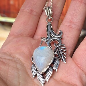 Op de afbeelding: Een zilveren ketting met een hanger met een grote, traanvormige maansteen, gezet in een gedetailleerd zilveren ontwerp. De hanger heeft ingewikkelde bloem- en bladmotieven.