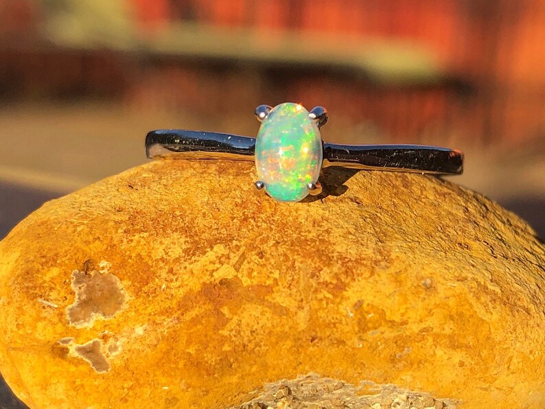 May include: A silver ring with a green opal gemstone set in a prong setting. The ring is resting on a yellow rock.