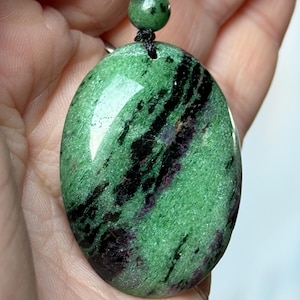 May include: Oval-shaped green pendant with black and maroon veining, suspended from a black cord necklace. The pendant has a polished, glossy surface. A small green bead accents the cord near the pendant.