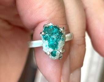 May include: A silver ring with a large, green, crystal-like stone set in the center. The stone is rough and has a natural, organic look.