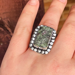 May include: A silver ring with a green and black stone set in a textured silver bezel. The ring is on a finger.