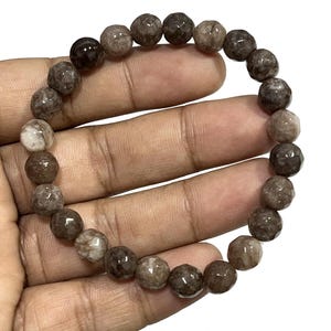 以下が含まれることがあります： 茶色と白の宝石のブレスレットで、ファセットカットのビーズが使用されています。ブレスレットは天然石で作られており、長さは約18cmです。