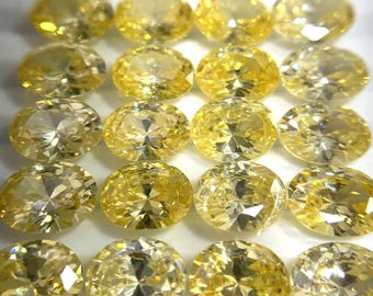 May include: A collection of 20 oval-shaped, faceted, yellow gemstones. The gemstones are arranged in rows on a white surface.