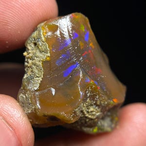 May include: A raw, unpolished opal gemstone with a translucent, amber-brown base colour. The stone features vibrant flashes of iridescent blue, green, and red play-of-colour across its smooth, glassy surface, contrasted by rough, textured, earthy edges.