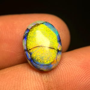 May include: An oval opal gemstone, displaying a striking array of colours. The centre of the opal is a shimmering gold, encircled by bands of blue and touches of orange. The stone is held between fingers, highlighting its natural beauty and complex patterns.